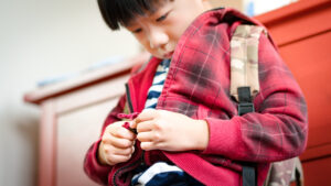 Young boy concentrating on zipping up his red jacket while wearing a backpack