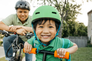 Young boy wearing a green helmet and shirt on a bicycle smiling at camera with his father in the background smiling and watching from a bike