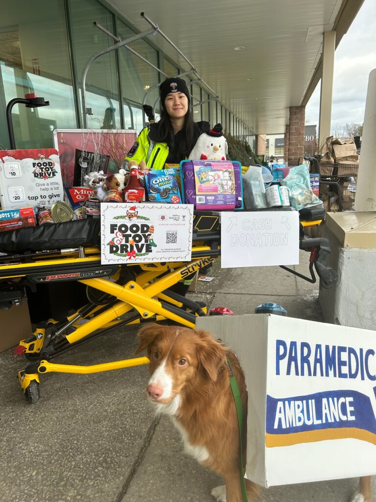 A paramedic smiles while displaying donated toys on a stretcher. A brown and white dog wearing an ambulance costume is in the foreground looking at the camera.